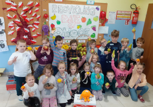 grupa dzieci pozuje do zdjęcia, w rękach mają owoce, za nimi plakat promujący akcję