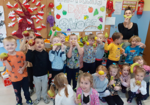 nauczycielka i grupa dzieci pozują do zdjęcia, w rękach mają owoce, za nimi plakat promujący akcję