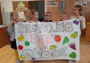 grupa dzieci trzyma w rękach duży plakat promujący akcję. Jest na nim napis recykling daje owoce oraz rysunki różnych owoców