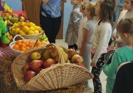 dzieci przy punkcie zbiórki baterii, słuchają nauczycielki, która rozmawia z nimi o owocach