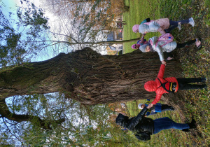 dzieci trzymają się za ręce i stoją wokół pnia drzewa, razem z nimi stoi pani