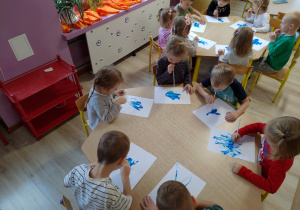 grupa dzieci siedzi przy stoliku, trzymają w ustach słomki, przez które dmuchają na niebieską farbkę, tworząc plamy