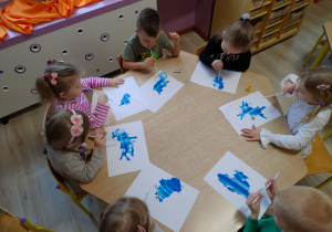 grupa dzieci siedzi przy stoliku, trzymają w ustach słomki, przez które dmuchają na niebieską farbkę, tworząc plamy