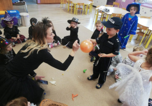 dzieci i nauczycielka przebrana za czarownicę siedzą w kręgu na dywanie, jeden z chłopców trzyma balon, który nauczycielka przebija szpilka