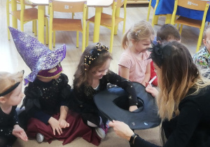 nauczycielka przebrana za czarownicę podaje dzieciom kapelusz, z którego losują wróżby
