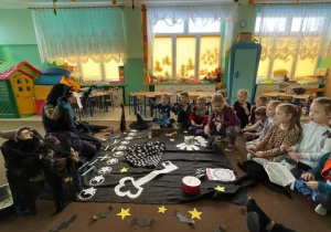 nauczycielka przebrana za czarownicę i grupa dzieci zgromadzeni w kręgu, przed nimi na dywanie leżą elementy dekoracji związane z andrzejkami