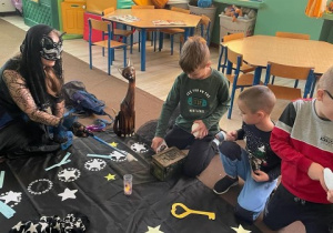 nauczycielka przebrana za czarownicę i grupa dzieci zgromadzeni w kręgu, przed nimi na dywanie leżą elementy dekoracji związane z andrzejkami