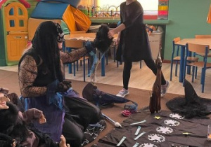 nauczycielka przebrana za czarownicę w dłoni trzyma czarną torebkę, z której jedna z dziewczynek losuje wróżbę