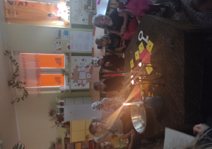 grupa dzieci siedzi wokół stolika, na nim stoją: świeczki misa z wodą i inne andrzejkowe atrybuty