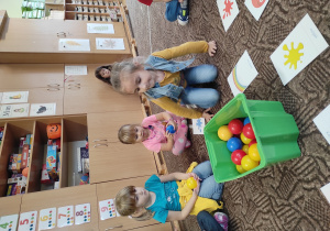 dzieci siedzą na podłodze , jedno z dzieci na środku, na podłodze stoi pudełko z kolorowymi piłkami