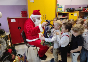 Mikołaj siedzi i rozdaje prezenty, przed nim stoi grupa dzieci