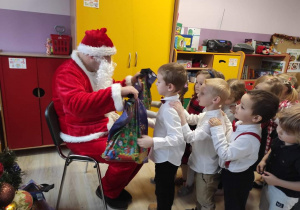Mikołaj siedzi i rozdaje prezenty, przed nim stoi grupa dzieci