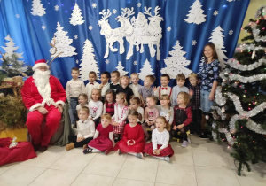 grupa dzieci ze swoimi paniami pozuje do zdjęcia z Mikołajem na tle świątecznej dekoracji