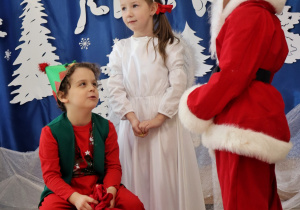 troje dzieci przebranych za: elfa, Mikołaja, anioła, na tle świątecznej dekoracji
