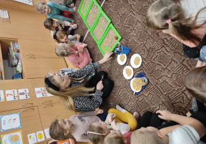 dzieci siedzą na podłodze, z nimi nauczycielka, przed nią leżą tace z mieszankami ziaren