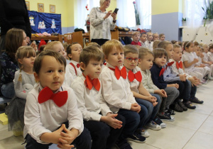 dzieci w białych koszulach i czerwonych muchach siedzą na ławeczce w szatni, czekają na występ
