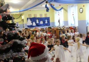 grupa dzieci tańczy w szatni, dziewczynki w białych sukienkach, obok siedzi Mikołaj