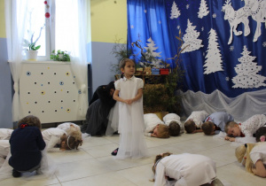 występ najmłodszej grupy, dziewczynka w białej sukience stoi, wokół niej dzieci leżą na podłodze