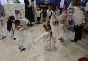 grupa dzieci odświętnie ubranych tańczy z białymi chustkami , obok jest ich nauczycielka