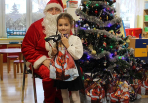Dziewczynka trzyma prezent, stoi obok Mikołaja, przy nich jest choinka