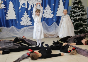 występ dzieci, jedna dziewczynka "Aniołek" stoi w środku, ma uniesione ręce, obok niej kilku chłopców "diabłów" leży na podłodze, obok dziewczynka przebrana za zimę