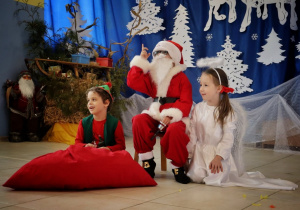 chłopiec "elf" i dziewczynka "aniołek" siedzą na podłodze, w środku Chłopiec "Mikołaj" siedzi na krzesełku