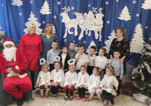 grupa dzieci ubranych odświętnie, nauczycielki, dyrektor i Mikołaj pozują do zdjęcia na tle świątecznej dekoracji