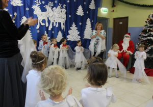 grupa dzieci ubranych odświętnie tańczy z białymi chusteczkami, obok stoi nauczycielka, na krześle siedzi Mikołaj