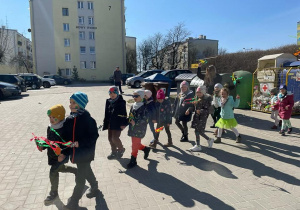 dzieci maszerują w barwnym korowodzie ulicami miasta, w rękach trzymają gaiki, instrumenty, na głowach mają wiosenne opaski