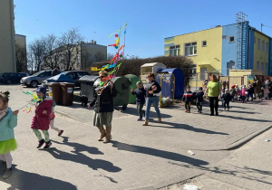 dzieci maszerują w barwnym korowodzie ulicami miasta, w rękach trzymają gaiki, instrumenty, na głowach mają wiosenne opaski