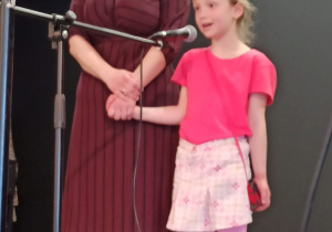 dziewczynka w różowym ubraniu mówi wiersz do mikrofonu, obok niej stoi nauczycielka
