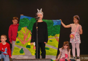 chłopiec recytuje wiersz na scenie, za nim 4 dzieci trzyma element scenografii