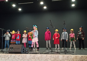 grupa dzieci występuje (śpiewa) na scenie, na głowach mają czapki z logo przedszkola