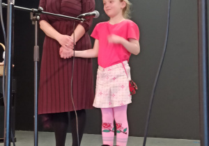 dziewczynka recytuje wiersz do mikrofonu, obok niej stoi nauczycielka