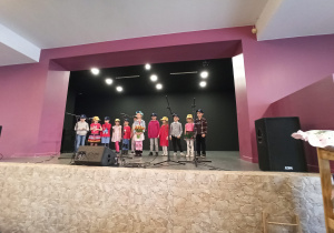 grupa dzieci występuje (śpiewa) na scenie, na głowach mają czapki z logo przedszkola