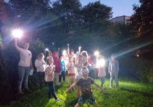 noc, dzieci i nauczycielka w przedszkolnym ogrodzie, w dłoniach trzymają latarki