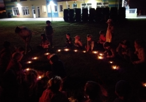 noc, grupa dzieci w przedszkolnym ogrodzie siedzi w kręgu, przy nich zapalone świeczki