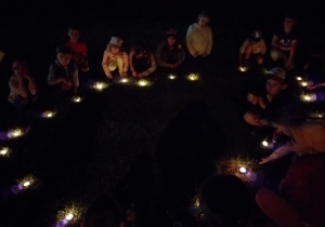 noc, grupa dzieci w przedszkolnym ogrodzie siedzi w kręgu, przy nich zapalone świeczki
