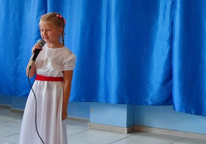 dziewczynka ubrana w białą suknię deklamuje wiersz, w tle dekoracja