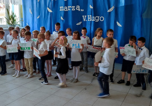 grupa dzieci ubranych galowo, w dłoniach trzymają tabliczki z wypisanymi własnymi marzeniami