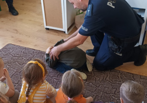 grupa dzieci siedzi na dywanie ,jedno z dzieci w pozycji "na żółwia" prezentuje jak się zachować podczas ataku psa , obok dziecka kuca policjant