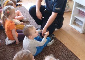 grupa dzieci siedzi na dywanie ,jedno z dzieci przymierza kajdanki, obok dziecka kuca policjant