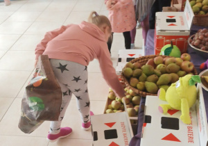 dziewczynka wrzuca zużyte baterie do specjalnego pojemnika przy niej stoją dzieci, obok leżą różne owoce