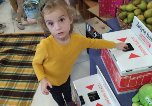 dziewczynka wrzuca zużyte baterie do specjalnego pojemnika, obok w koszach znajdują się owoce