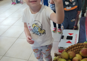 dziewczynka trzyma w dłoni jabłko, obok znajdują się owoce w koszach oraz pojemniki na zużyte baterie