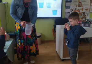 Chłopiec pije wodę ze szklanki. Pani dietetyk stoi obok, w ręku trzyma dzbanek z wodą.
