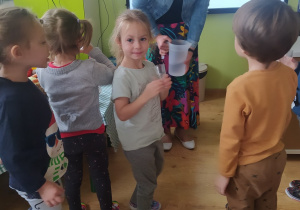 Dzieci piją wodę, jedna z dziewczynek pozuje do zdjęcia. Pani dietetyk stoi obok, w ręku trzyma dzbanek z wodą.
