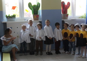 Grupa dzieci w białych pelerynach i strojach misiów stoi przed publicznością, nauczycielka siedzi obok na ławce.