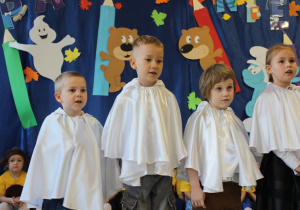 Grupa dzieci w białych pelerynach stoi przed publicznością i śpiewa piosenkę.