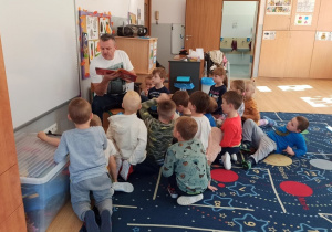 Tata Filipa siedzi na krzesełku i czyta dzieciom bajkę. Dzieci siedzą przed nim na dywanie, z zainteresowaniem słuchają utworu.